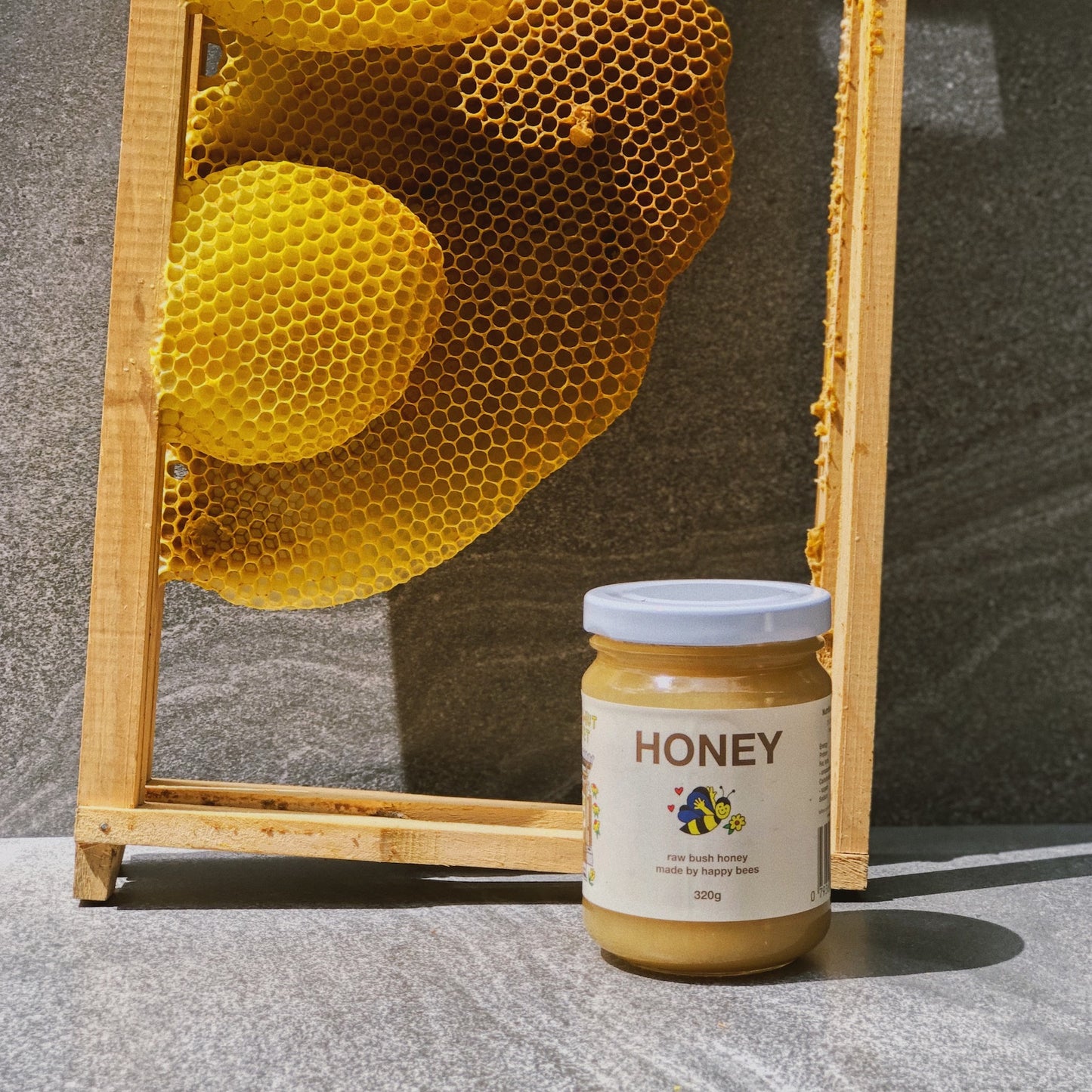 A 320g jar of Friday Hut Market Raw Bush Honey. Frames from a beehive are in the background with golden beeswax honeycomb.