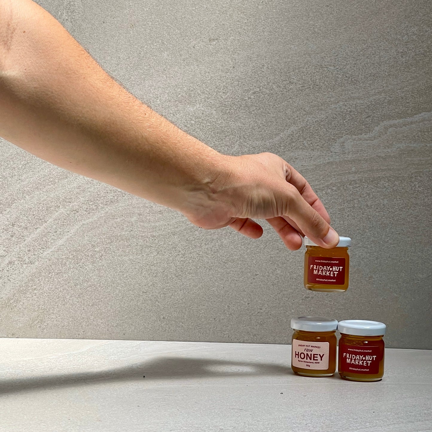 A hand is placing a 50g Sample Jar of Friday Hut Market Raw Bush Honey on top of two others.