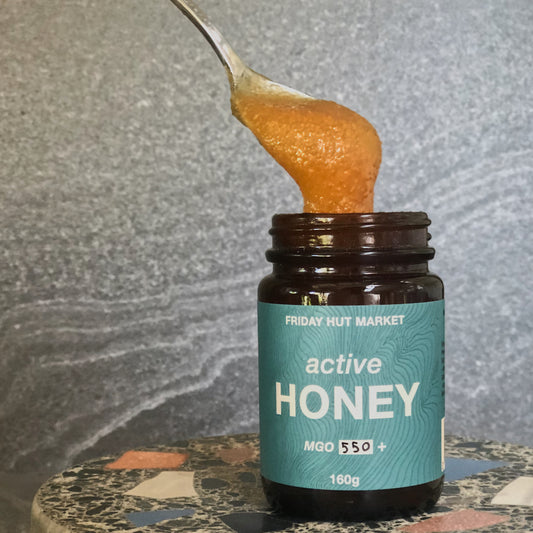 A spoonful of thick golden Friday Hut Market Active Jellybush Manuka honey is being taken from a medicinal looking amber jar.