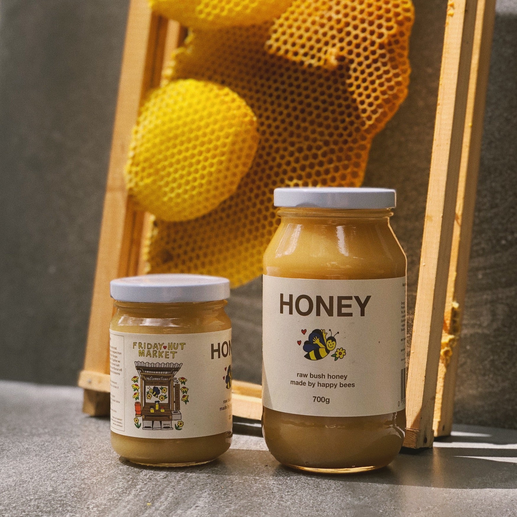 320g and 700g jars of Friday Hut Market Raw Bush Honey, made by happy bees. Frames from a beehive are in the background with golden beeswax honeycomb.