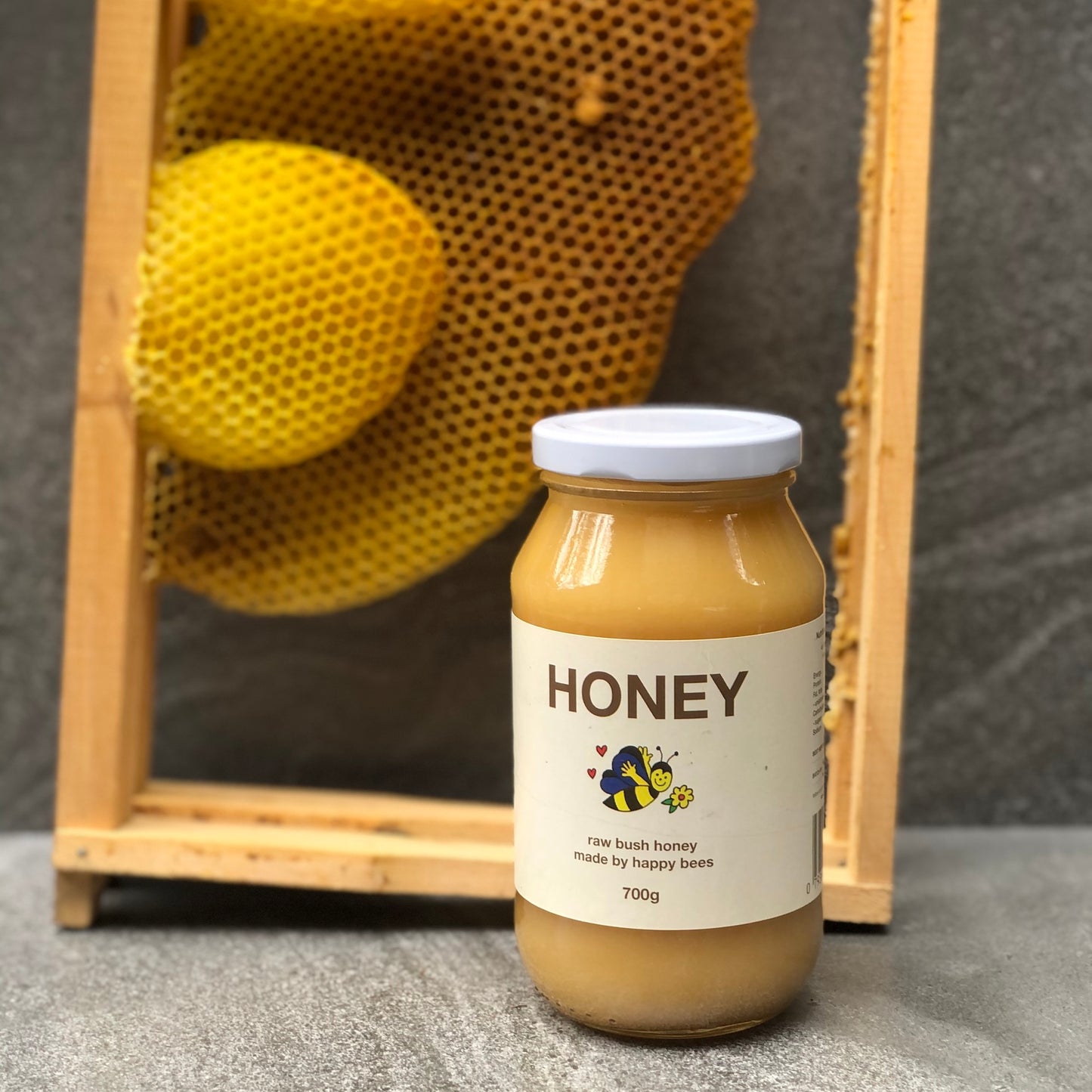 A 700g jar of Friday Hut Market Raw Bush Honey. Frames from a beehive are in the background with golden beeswax honeycomb.